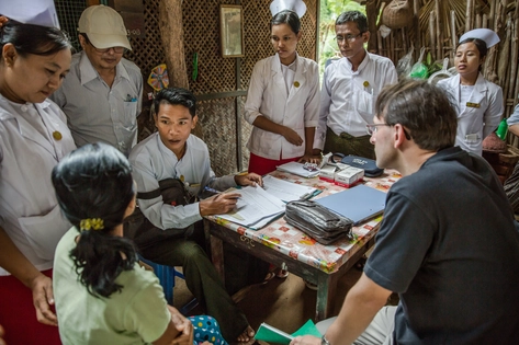 Foto de pessoas reúnidas ao redor de uma mesa com estampa colorida. O local é interno, com paredes trançadas de fibra e madeira. O círculo é composto de oito pessoas, uma delas vestida de branco direciona o olhar para uma mulher de costas que veste uma roupa verde claro. Ele tem papéis e faz anotações. Ao redor, a equipe veste jaleco branco, chapéu de enfermagem, alguns boné. 